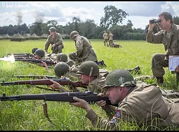 Soldiers in uniform aiming their rifles at a gun range, one has fired, there is a soldier with a par of binoculars looking down range.